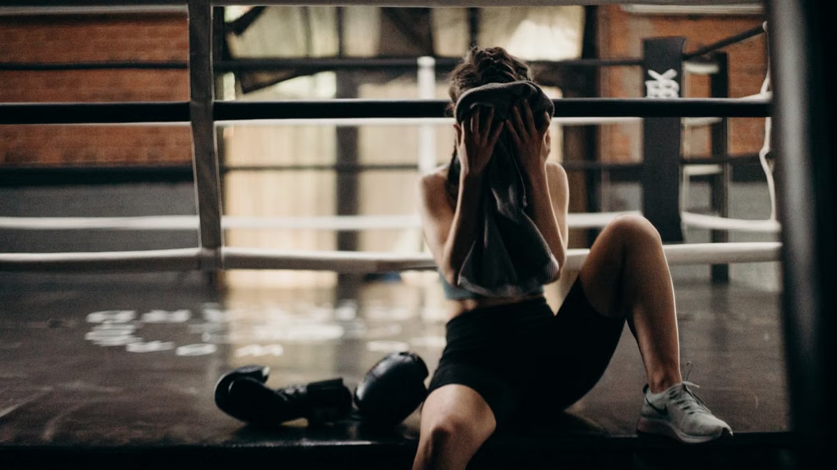 A female boxer sitting in a gym ring, resting and wiping sweat with a towel after a workout.